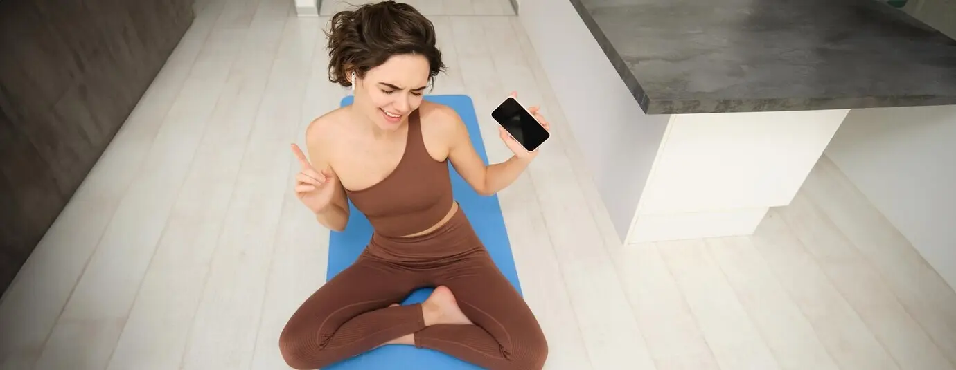 Танцующая брюнетка сидит на коврике для йоги с наушниками в ушах и слушает музыку во время тренировки.